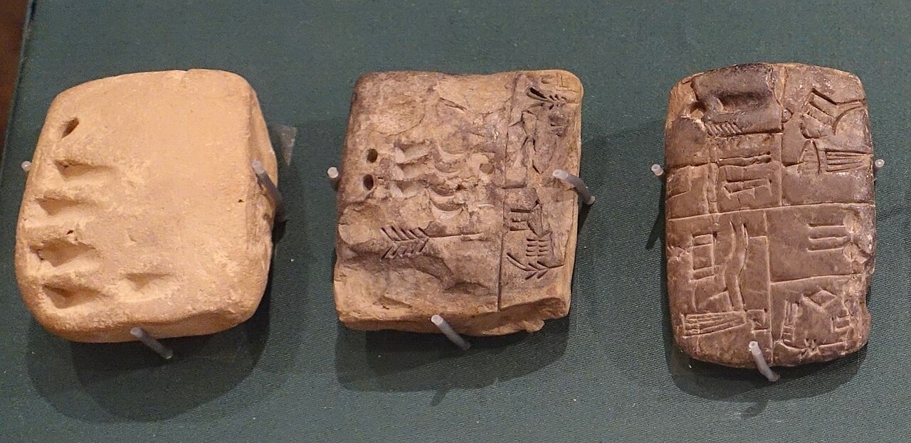 Three small clay tablets side by side. The left one bears only impressed numerical marks. The center one mixes numbers with simple pictures. The right one carries early proto-cuneiform signs.